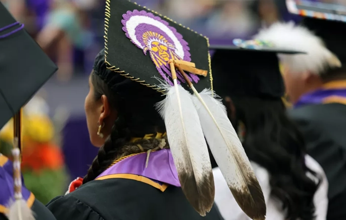 A close-up view of Native American graduates in caps and gowns at a graduation ceremony. The central graduate wears a decorated cap featuring a detailed beaded design of a Native figure in purple, white, and gold, bordered with gold thread and adorned with two large white feathers hanging down the back. Other graduates nearby also wear traditional regalia elements with their academic attire. The scene conveys pride, cultural identity, and academic achievement.
