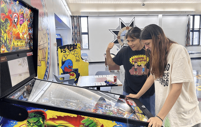 Two young men are playing an Avengers-themed pinball machine in a brightly lit indoor recreational area.