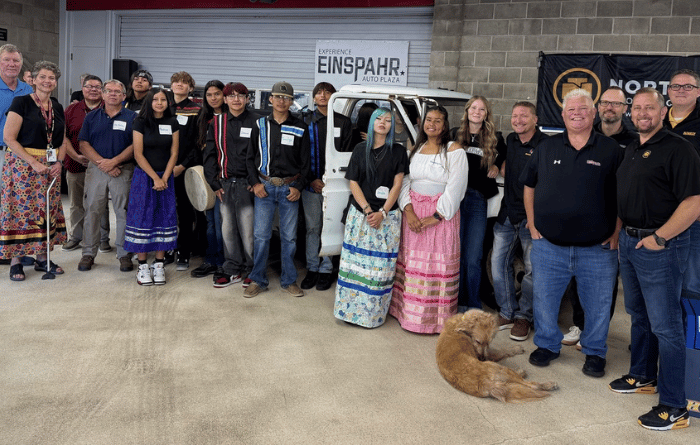 A group poses outside an auto shop beside a white utility vehicle, with a dog lying in front.