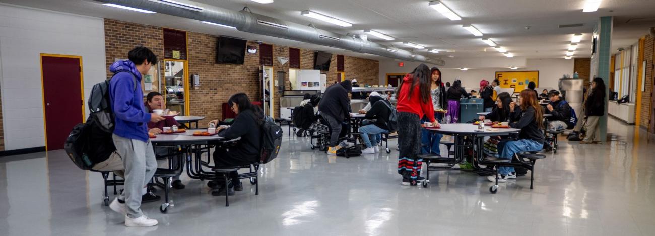 High school students enjoy lunch together in the bustling cafeteria, surrounded by tables and food trays.