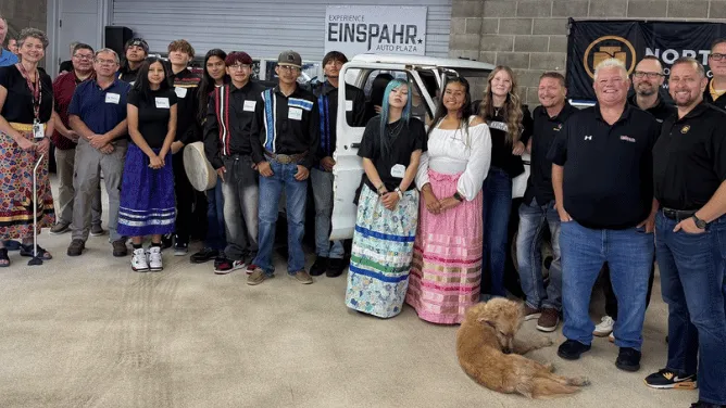 A group poses outside an auto shop beside a white utility vehicle, with a dog lying in front.
