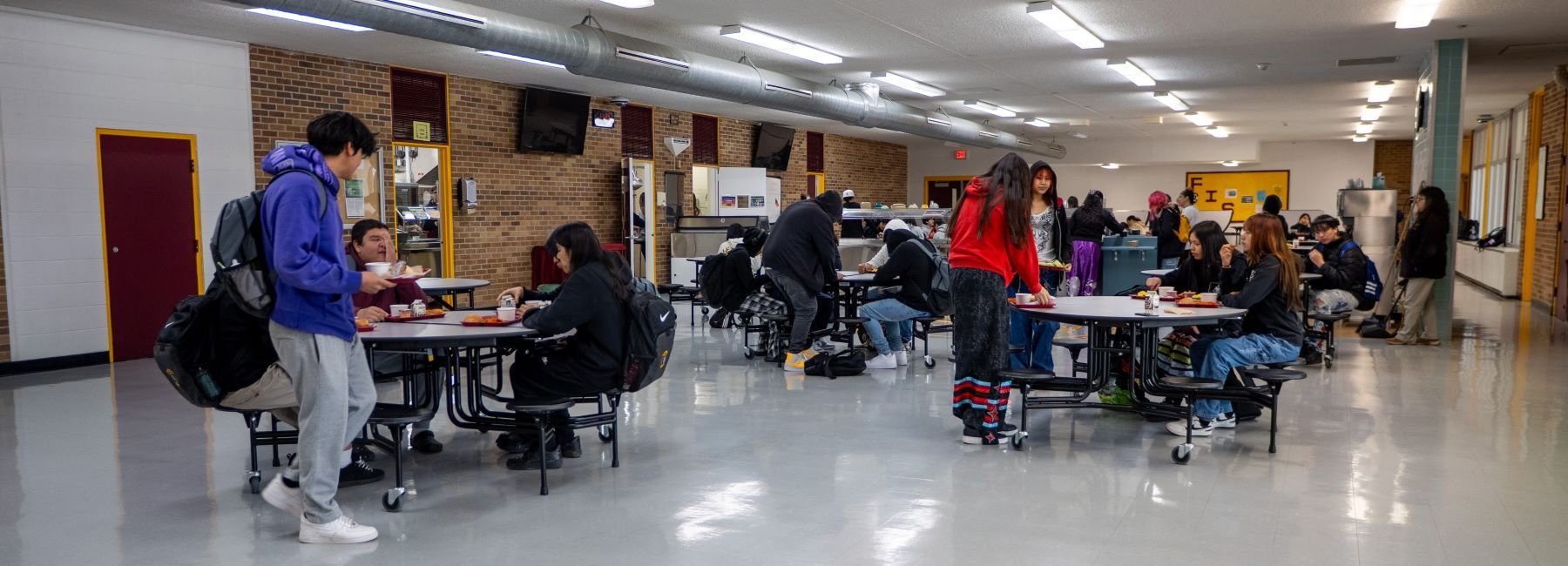 High school students enjoy lunch together in the bustling cafeteria, surrounded by tables and food trays.