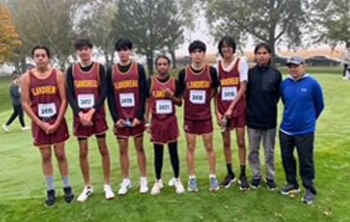 A boys cross country team stands in a line on a grassy field, wearing matching maroon uniforms with race bibs, alongside two adult coaches. Trees and other runners are visible in the background at a meet location.