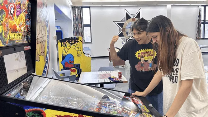 Two young men are playing an Avengers-themed pinball machine in a brightly lit indoor recreational area.